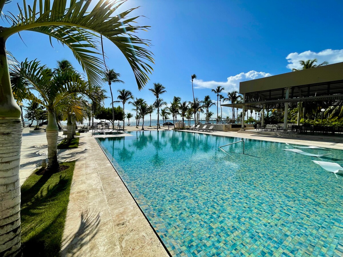 A large swimming pool with clear blue water reflects the sky, surrounded by tall palm trees and lounge chairs. The pool area is illuminated by sunlight, creating a serene outdoor space that opens to a view of the ocean.