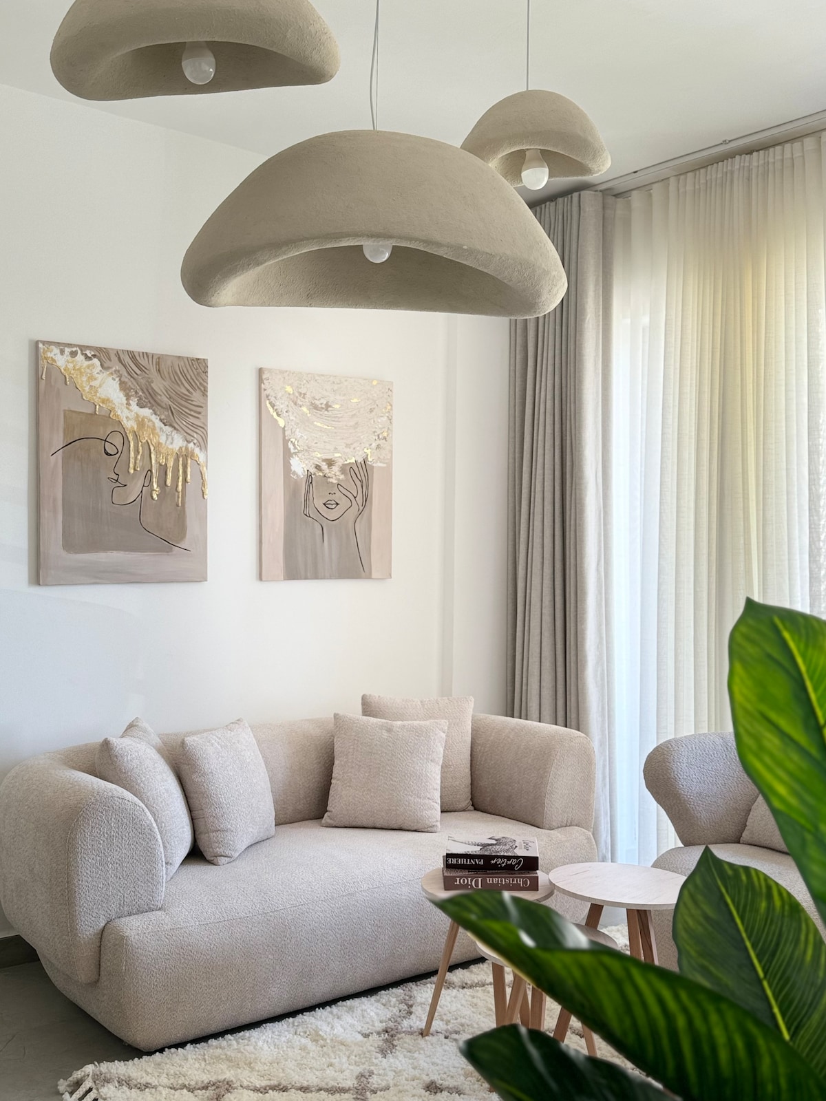 A light and airy living space is featured, with a soft beige sofa adorned with decorative pillows. The walls display two artistic pieces, while soft light filters through sheer curtains. A small round table sits nearby, accompanied by a potted plant, enhancing the cozy atmosphere.