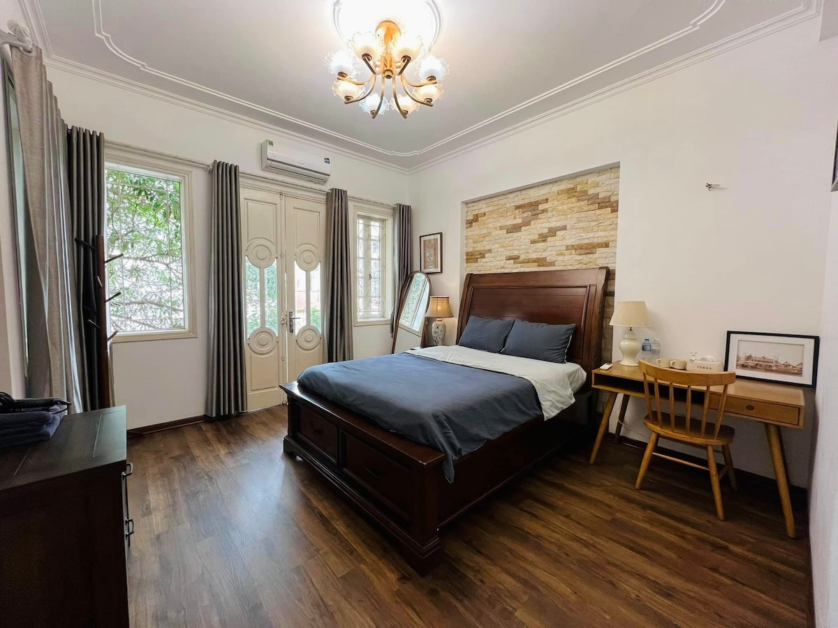 A spacious bedroom features a king-sized bed adorned with soft linens. Natural light enters through multiple windows, enhancing the wooden flooring and light-colored walls. A work desk and chair are positioned against one wall, while a chandelier adds an elegant touch to the ceiling.