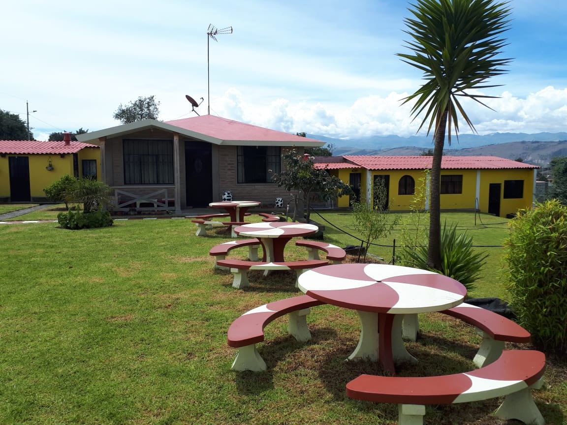 A lush green lawn features several circular picnic tables with red and white patterns. The tables are surrounded by well-maintained gardens, and the backdrop includes a charming house with a red roof and yellow walls, complemented by distant mountains under a clear blue sky.