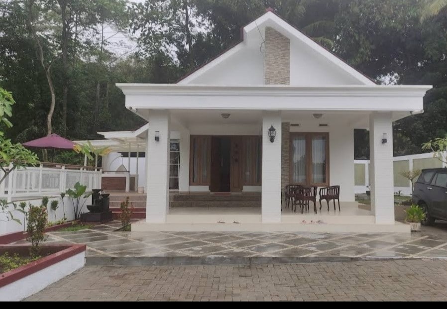 A serene exterior view of a white house featuring a covered porch and large windows. A paved driveway leads to the entrance, which is flanked by lush greenery and a shaded seating area. There is also a small outdoor dining set visible to the side.