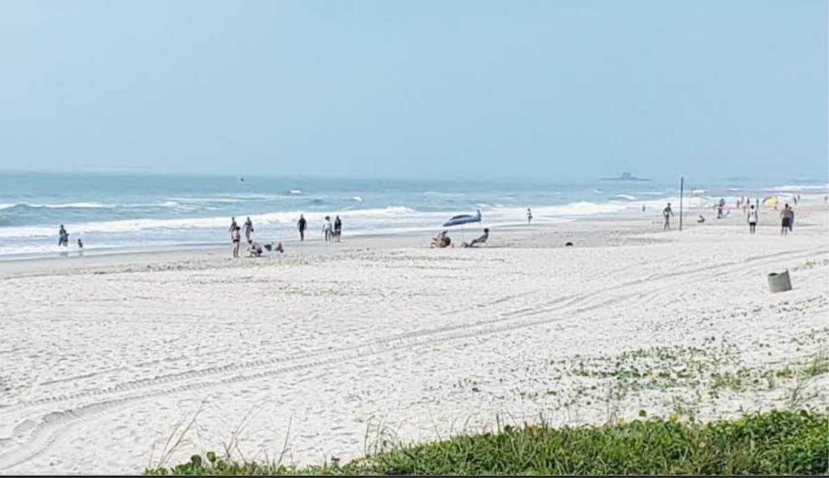 A sandy beach stretches along the coastline, with gentle waves lapping at the shore. Several individuals can be seen enjoying the beach, while distant boats float on the water. A clear sky enhances the serene coastal landscape.