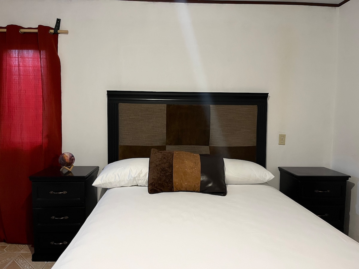 A spacious bed is positioned against a simple headboard, complemented by decorative pillows in various colors. Nightstands with drawers flank each side of the bed. A window covered with sheer curtains allows soft light to enter, while a red curtain adds a touch of color to the room.