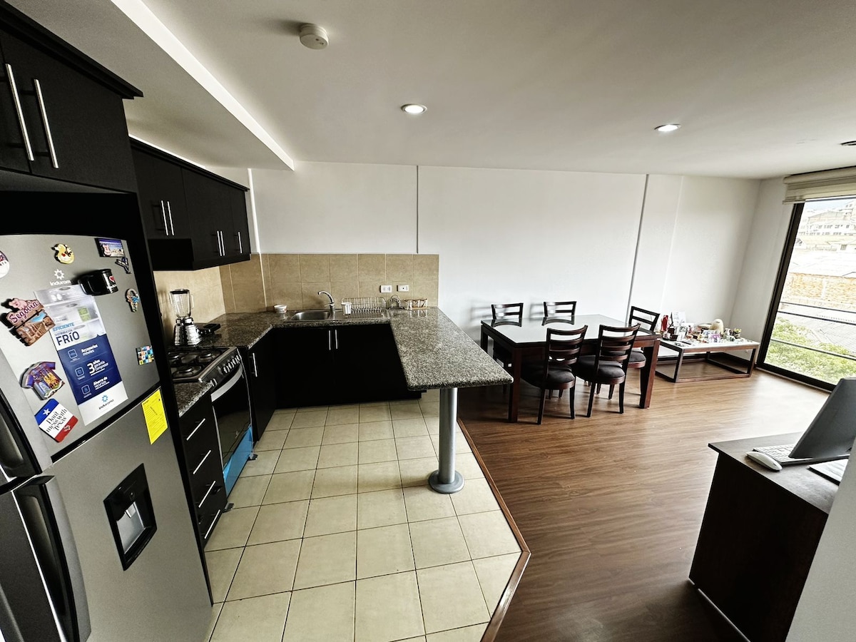 A modern kitchen and dining area are visible, featuring dark cabinetry and granite countertops. A stainless steel refrigerator and stove are included. A dining table with several chairs is positioned beside large windows, allowing natural light to fill the space. The floor combines tiles and wood textures.