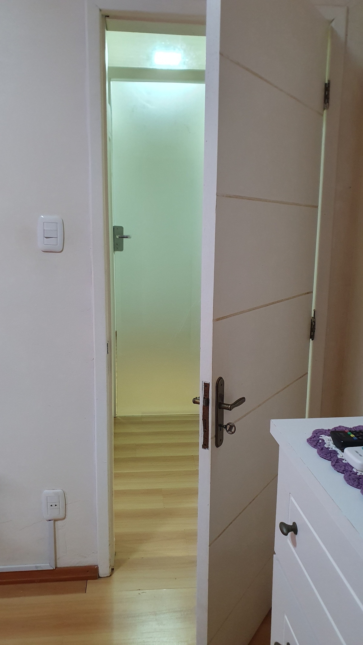 A partially open door reveals a softly illuminated hallway beyond. The light-colored wood floor extends into the distance, and a hint of a frosted glass panel can be seen in the doorframe, promoting a sense of privacy while inviting curiosity.