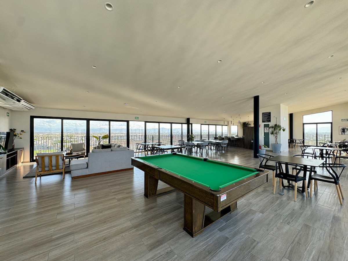 A spacious rooftop area features large windows offering panoramic views. A pool table is positioned prominently in the center, surrounded by various seating arrangements including tables and chairs. Soft natural light enhances the neutral tones of the flooring and walls.
