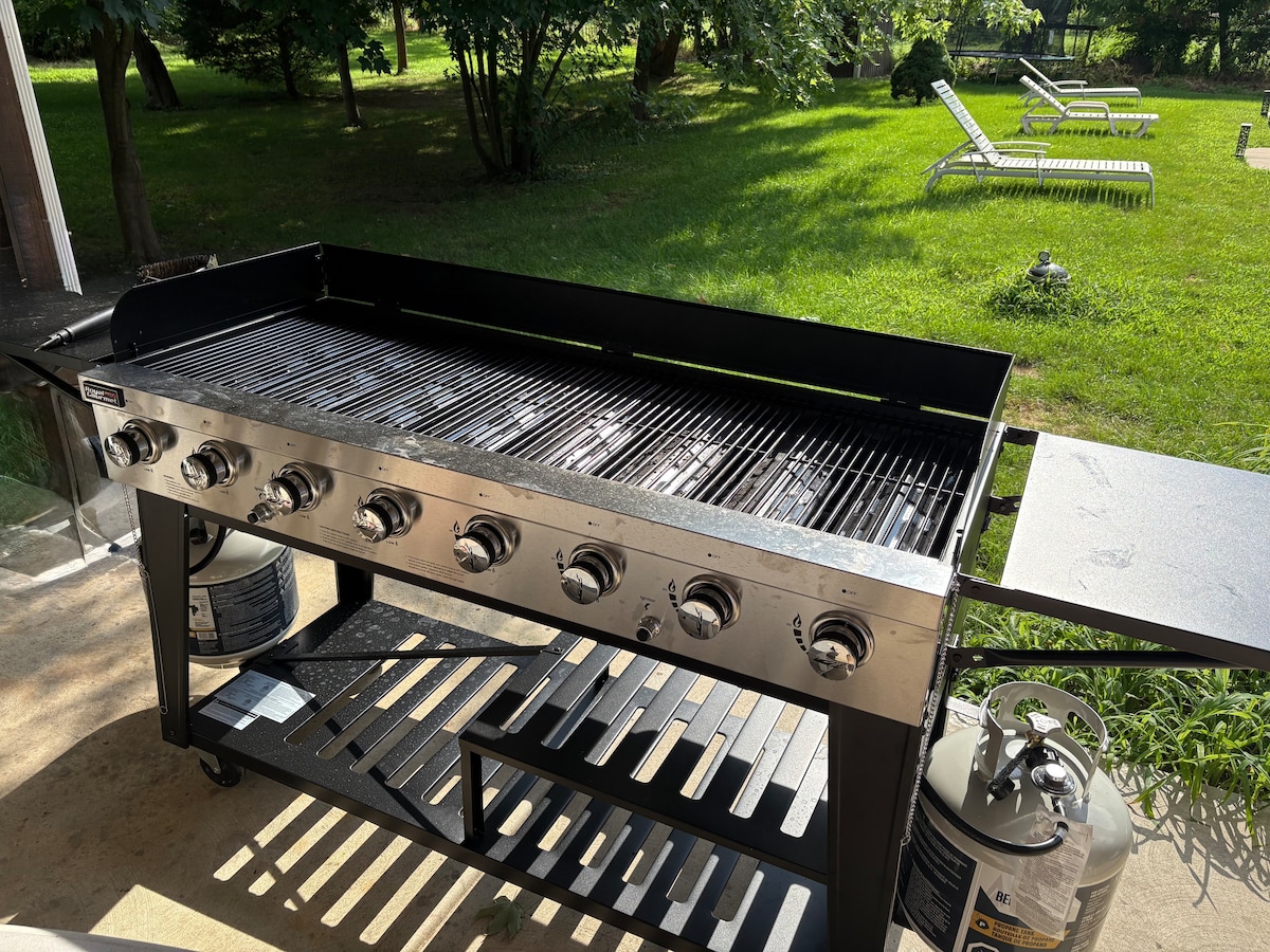 A large outdoor grill is positioned on a patio, featuring multiple burners and a sturdy cart for storage. Green grass and lounge chairs are visible in the background, contributing to a relaxing outdoor environment.
