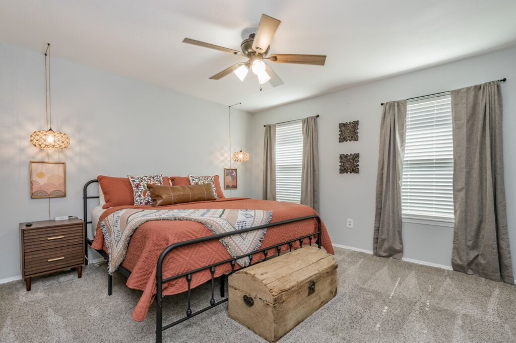 A spacious bedroom is presented with soft grey walls and a cozy bed dressed in warm-toned bedding. Two large windows allow natural light to fill the room, while pairs of decorative light fixtures hang above the bed. A decorative trunk sits at the foot of the bed.
