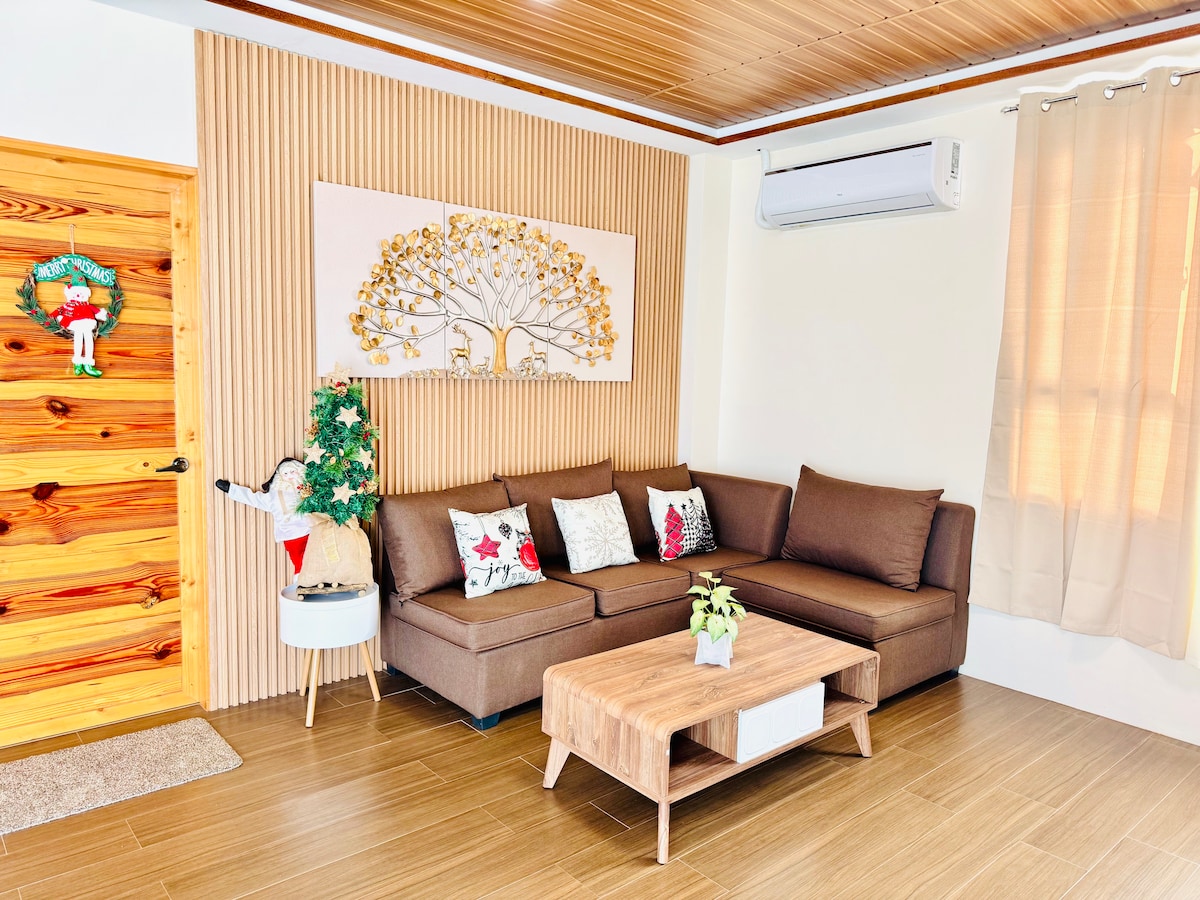 A spacious living area features a brown sectional sofa adorned with decorative pillows. A wooden coffee table sits in the center, complemented by a small potted plant. Natural light filters through sheer curtains, illuminating the textured wooden walls and decorative wall art.