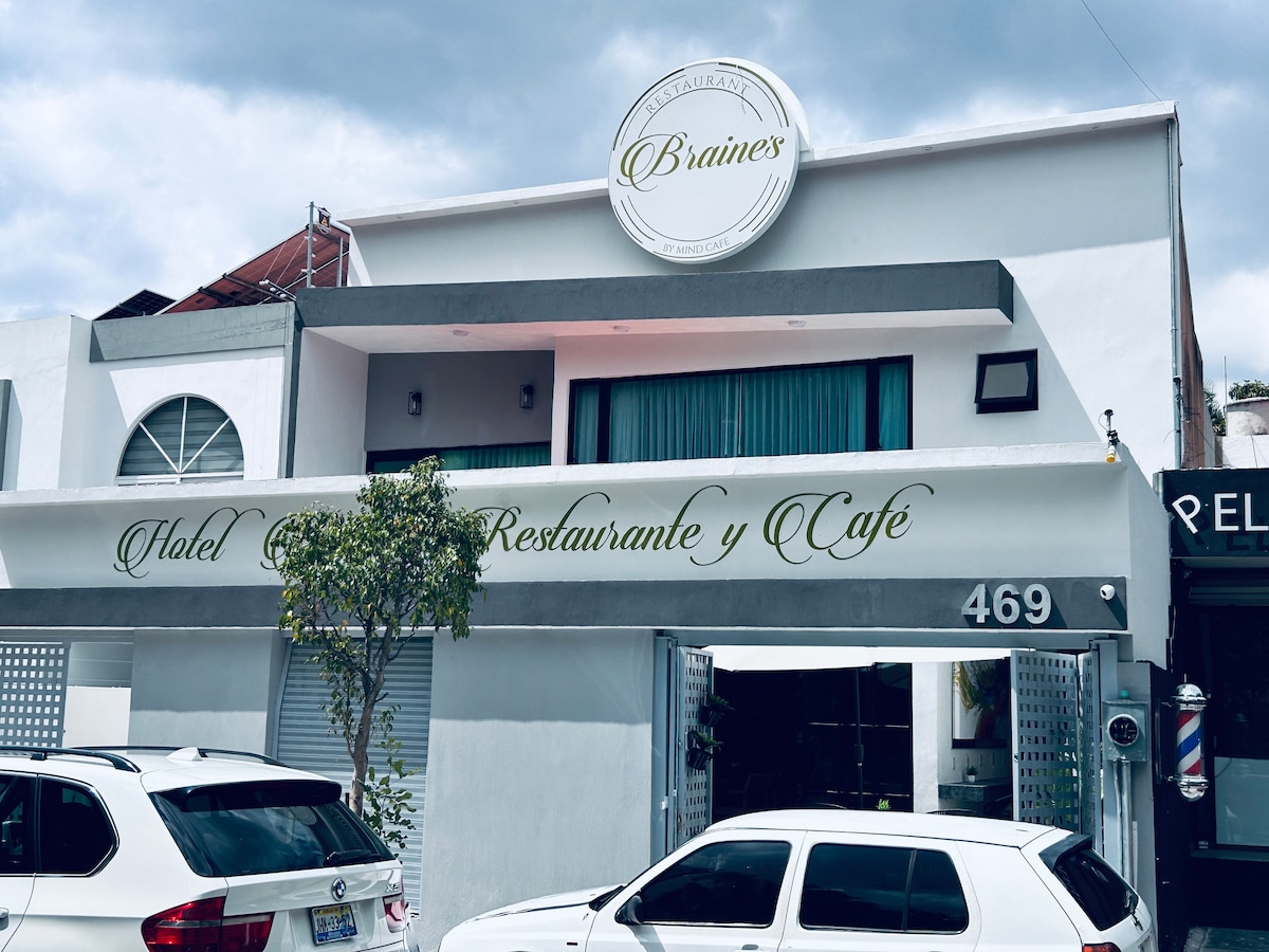 The exterior of a modern boutique hotel is highlighted, featuring a clean white facade with a prominent sign displaying the hotel's name. Large windows allow natural light into the interior, and a small tree adds greenery to the entrance. Parking spaces are visible in front.