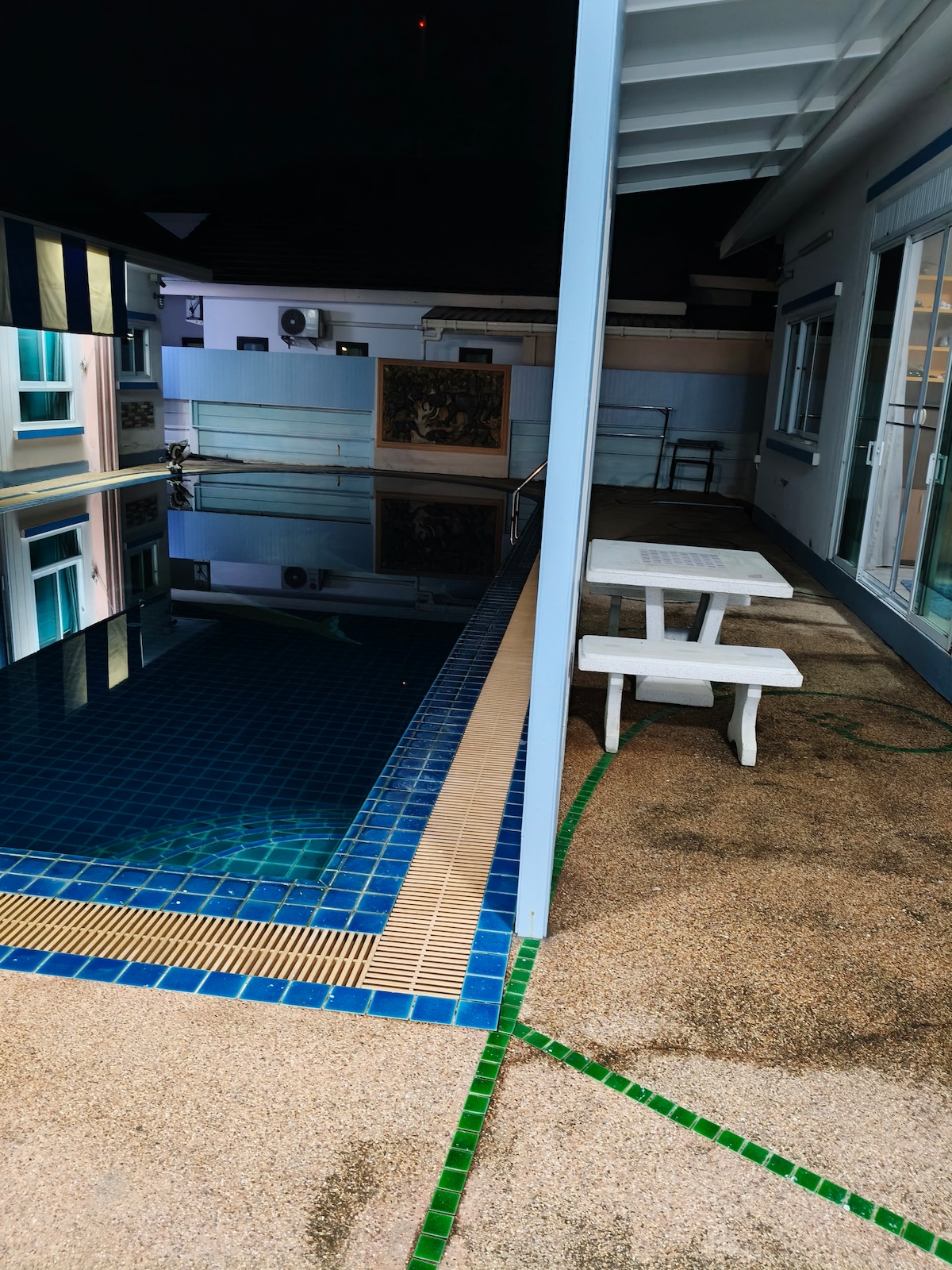 A shared outdoor space features a swimming pool with a blue tiled edge, reflecting surrounding lights. Adjacent to the pool, a white picnic table offers seating, set against a backdrop of the house's exterior. The area is well-lit, creating a welcoming ambiance.
