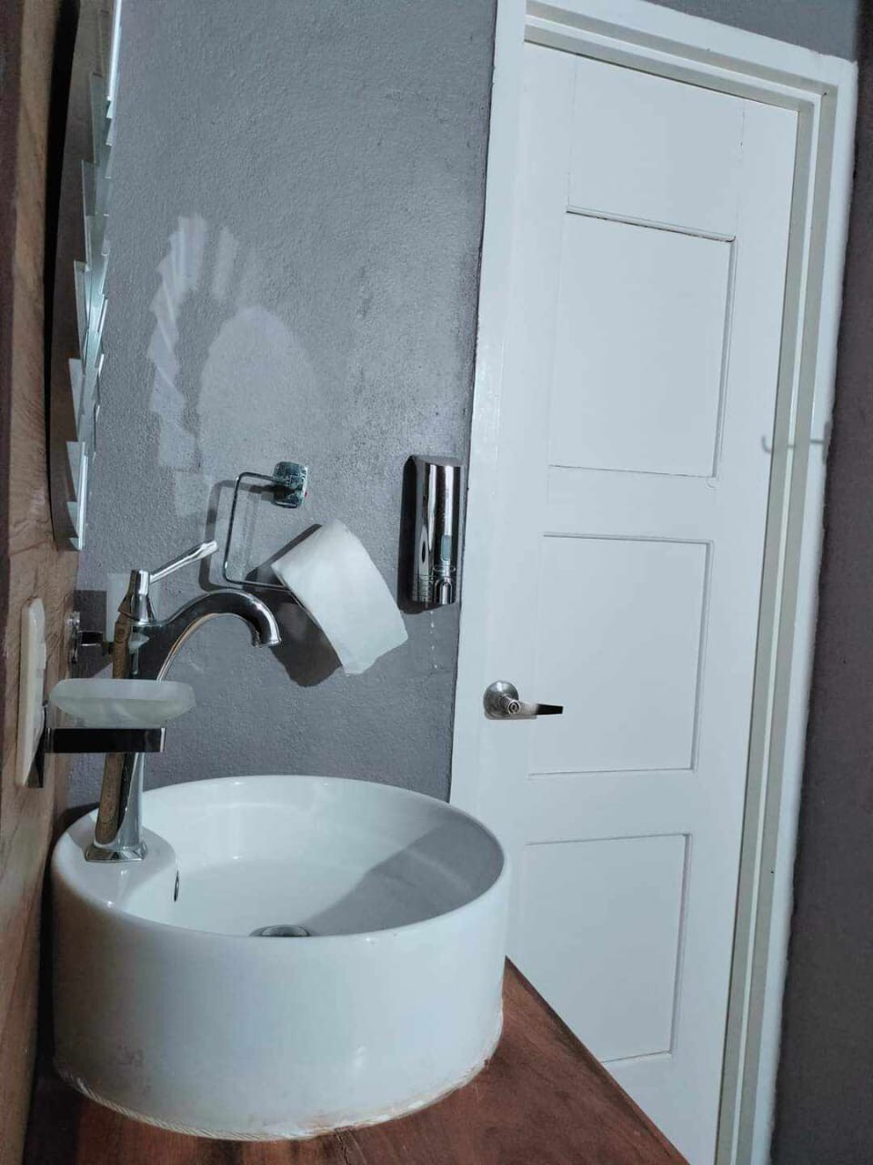 A modern bathroom sink is mounted on a wooden countertop, featuring a minimalistic round basin. A sleek faucet stands beside it, and a soap dispenser is positioned nearby. The wall is painted in a muted gray tone, and a closed white door is visible in the background.