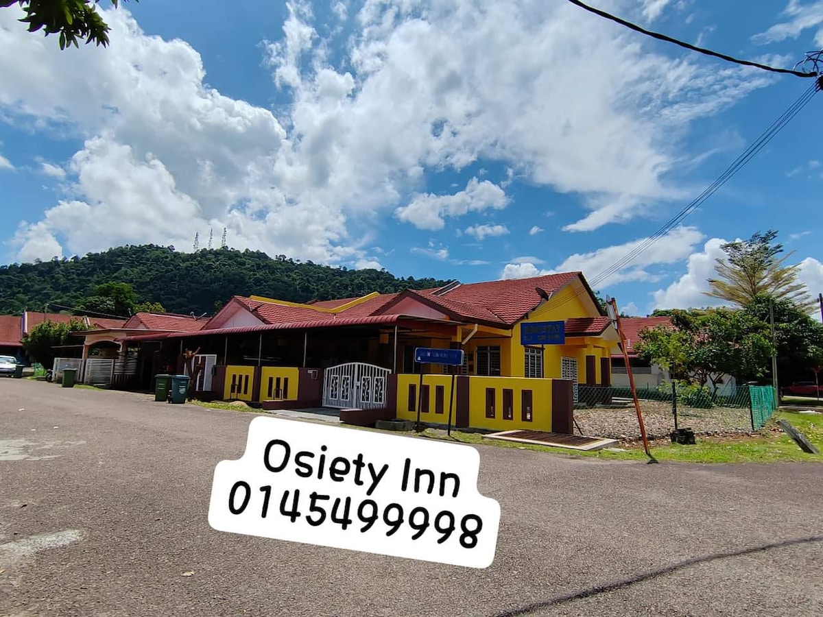 A single-story building is visible, featuring a yellow façade contrasting with red roofing. The surrounding area includes lush greenery and clear skies with scattered clouds, offering a serene backdrop. A sign prominently displays the name of the inn, contributing to the welcoming atmosphere.