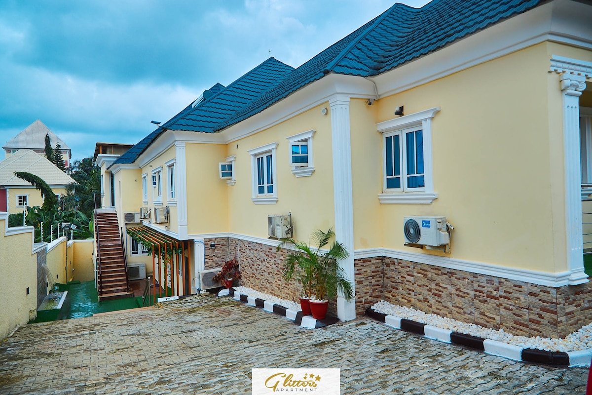 The exterior of the building is presented with a fresh color scheme. Air conditioning units are installed, ensuring comfort. Decorative stone accents are visible along the foundation. A pathway leads towards the entrance, flanked by greenery and subtle decorative elements.