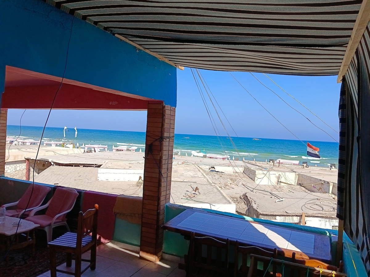 An open balcony provides a view of the beach, showcasing clear blue waters under a bright sky. Soft seating is visible, framed by colorful walls and striped awnings. A table invites relaxation while beach activity is seen in the distance, enhancing the coastal atmosphere.