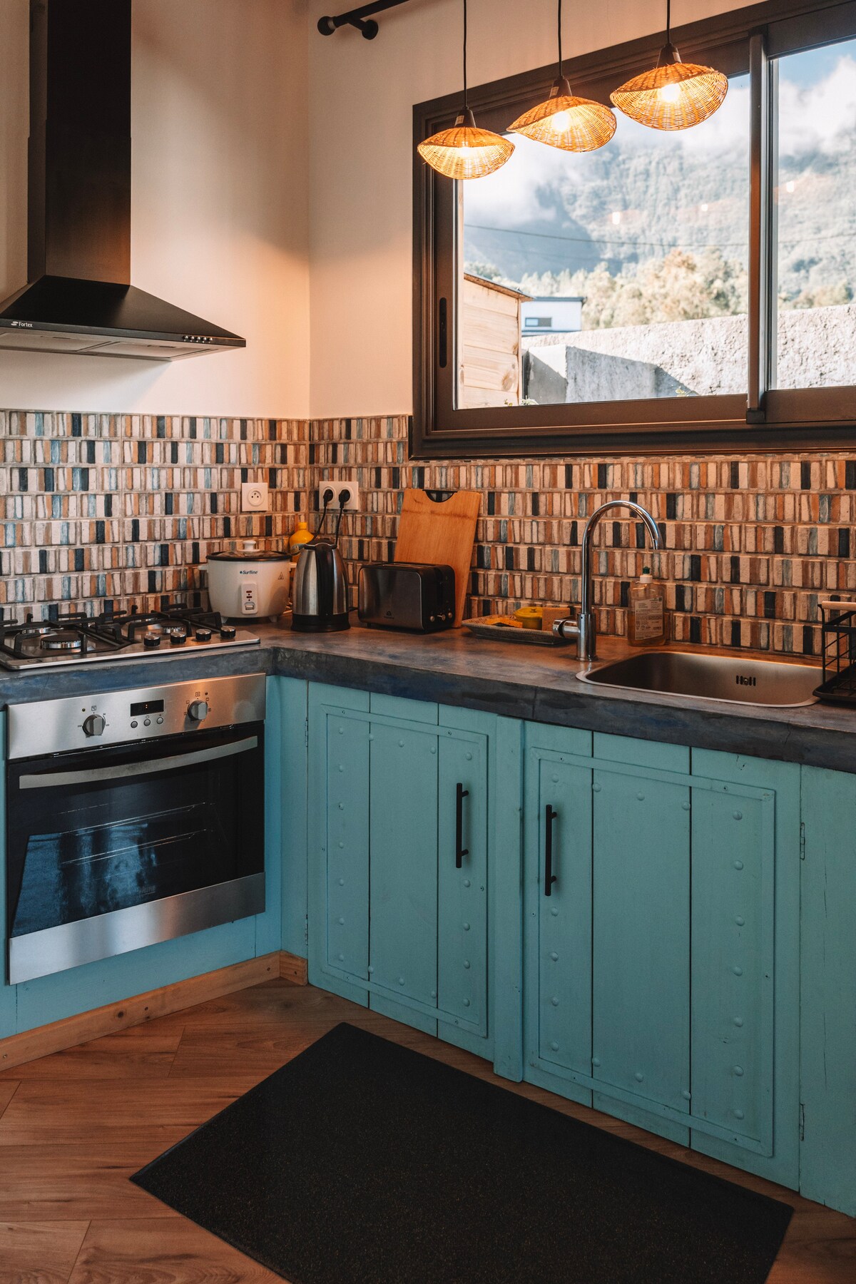 A well-equipped kitchen is presented with light blue cabinetry and modern appliances. A gas stove, oven, and stylish overhead lights are visible. The sink area features a sleek faucet, and a window offers natural light while showcasing a scenic view outside.