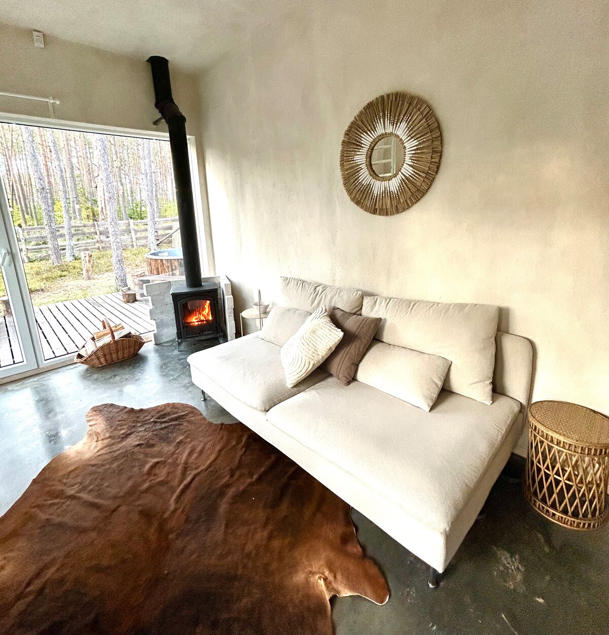 A comfortable sofa is positioned in a warm living space, with soft cushions arranged casually. A cozy wood-burning stove adds warmth, while large glass doors provide views of the surrounding pine forest and direct access to the outdoor terrace.