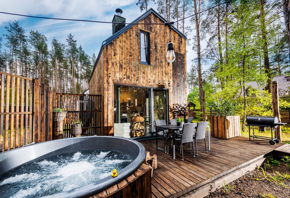The exterior of the wooden house showcases a spacious wooden terrace, featuring a hot tub and a barbecue grill. A dining set with chairs is positioned near the hot tub, and the surrounding forest provides a natural backdrop, enhancing the overall relaxing atmosphere.