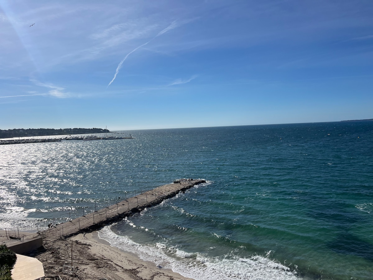A panoramic view of the sparkling blue sea is captured, with gentle waves lapping against a rocky jetty. The horizon merges with a clear sky, creating a serene atmosphere. The coastline is visible in the distance, enhancing the tranquil setting.