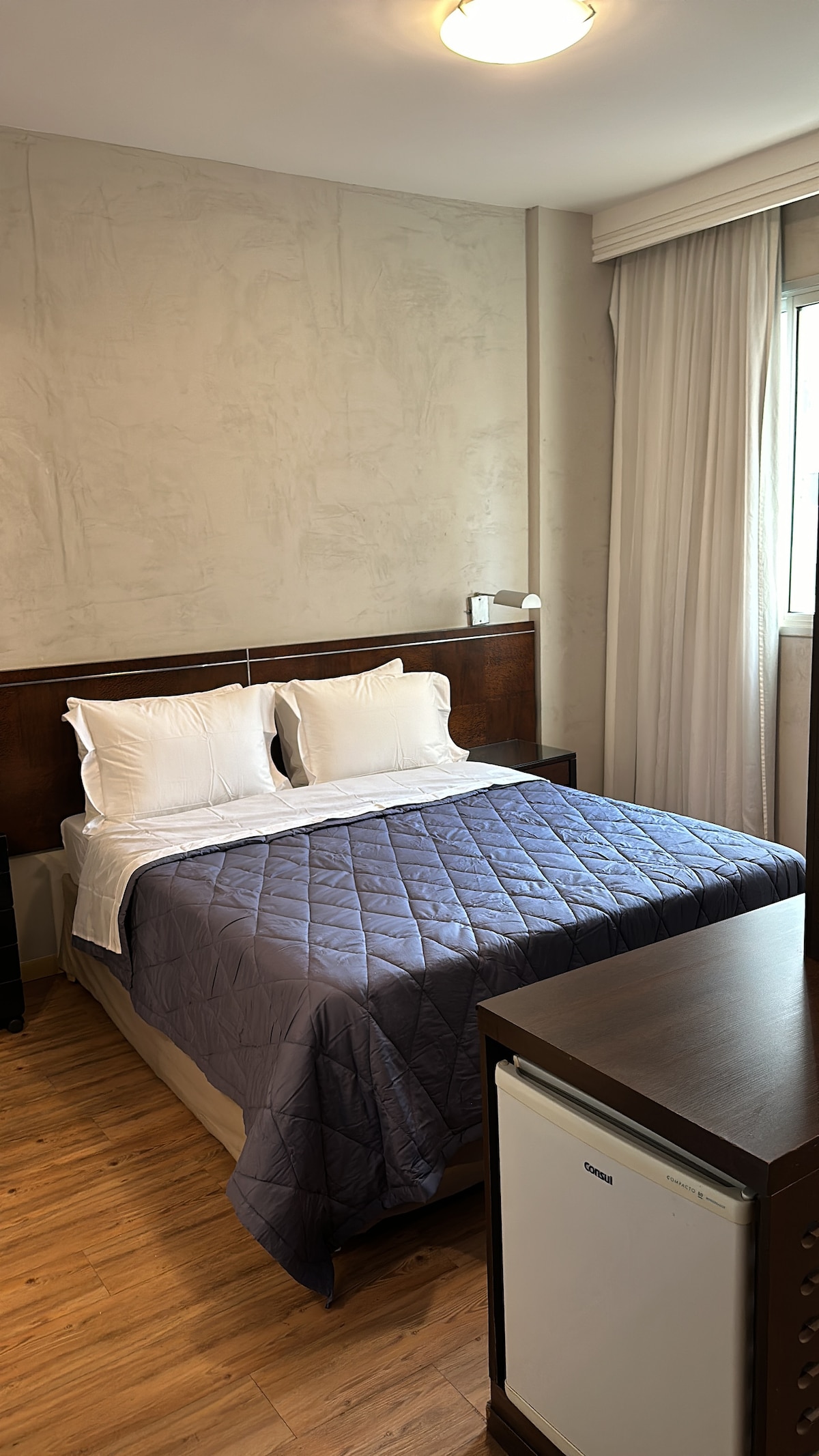 A well-appointed bedroom features a spacious bed with clean linens and a quilted coverlet. Natural light pours in through a window dressed with sheer curtains. A small refrigerator is positioned beside the bed, complementing the wooden furnishings and warm flooring.