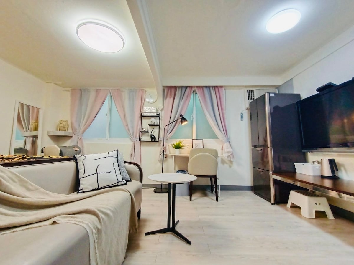 A modern living space features a comfortable sofa draped with a light blanket. A small, round table sits nearby, accompanied by a chair. Soft pastel curtains frame two windows, with a floor lamp illuminating the area. A refrigerator and a television are positioned against the wall.