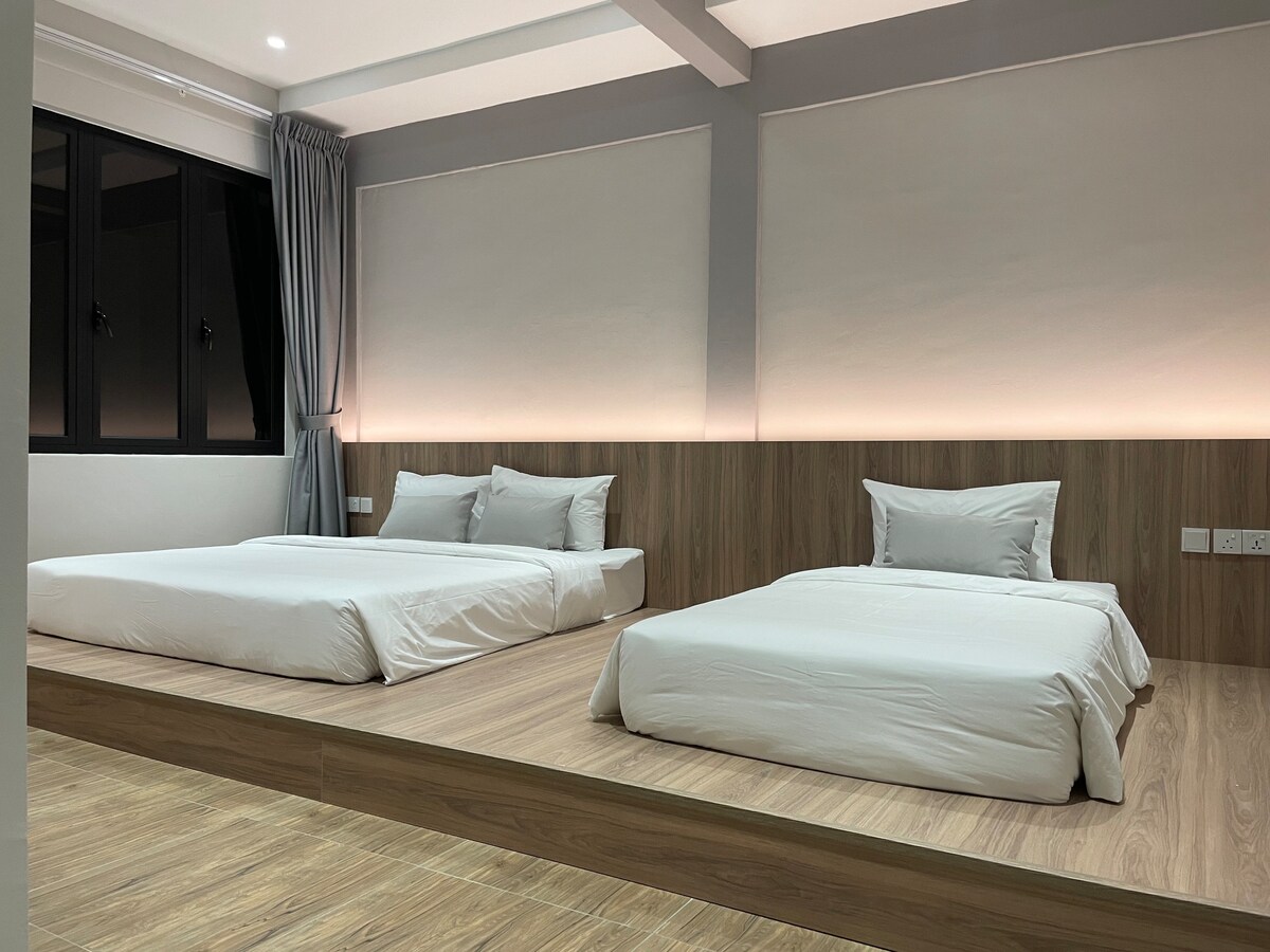 The sleeping area features two spacious beds arranged on a raised wooden platform. Soft natural light filters in through large windows, complemented by light-colored walls and subtle lighting along the base of the beds, creating a calming ambiance.