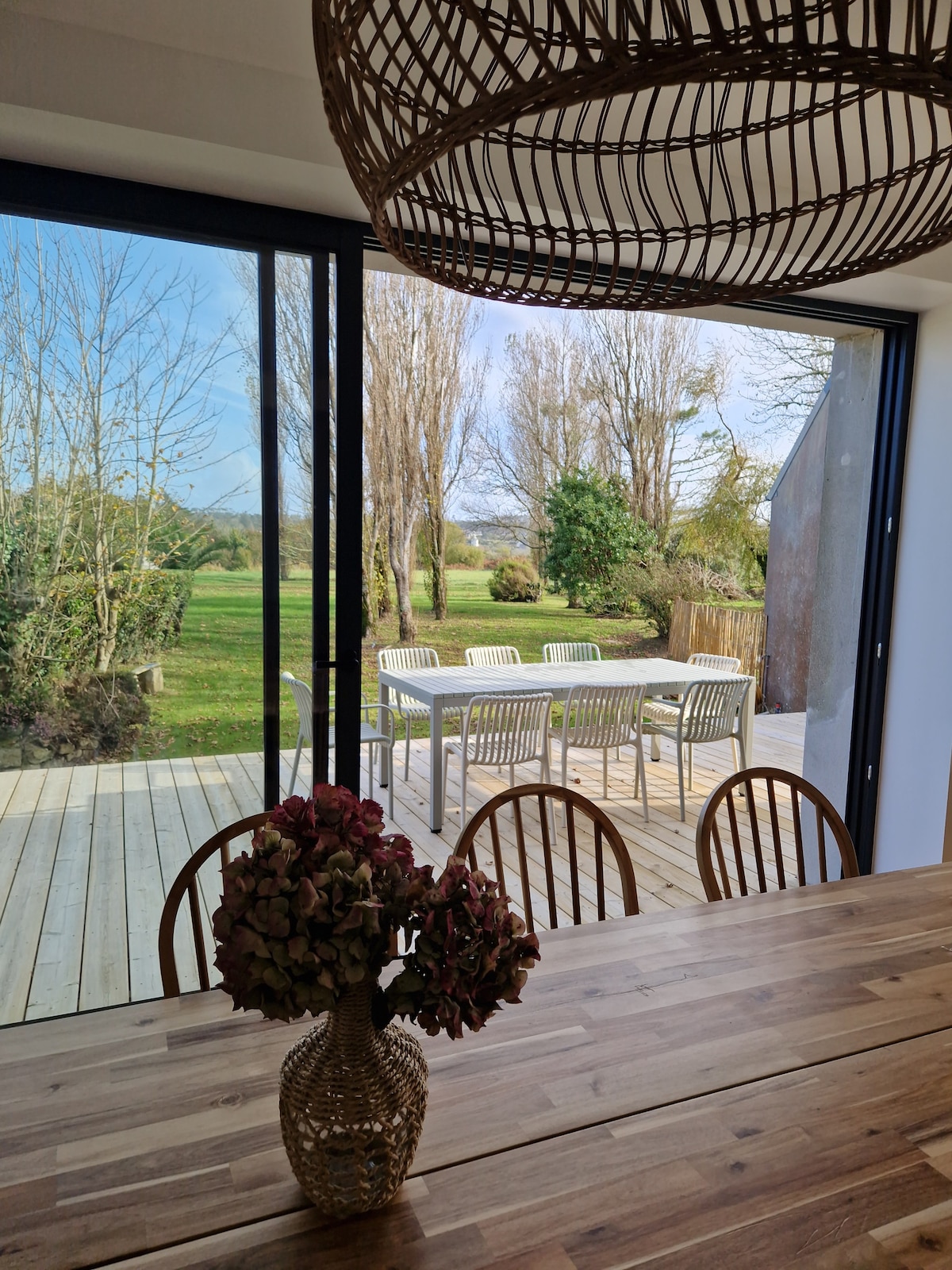 A spacious dining area features a wooden table surrounded by chairs, positioned near large glass doors. Outside, a wooden deck is visible, showcasing a dining set for six. Lush greenery and tall trees frame the serene outdoor space.