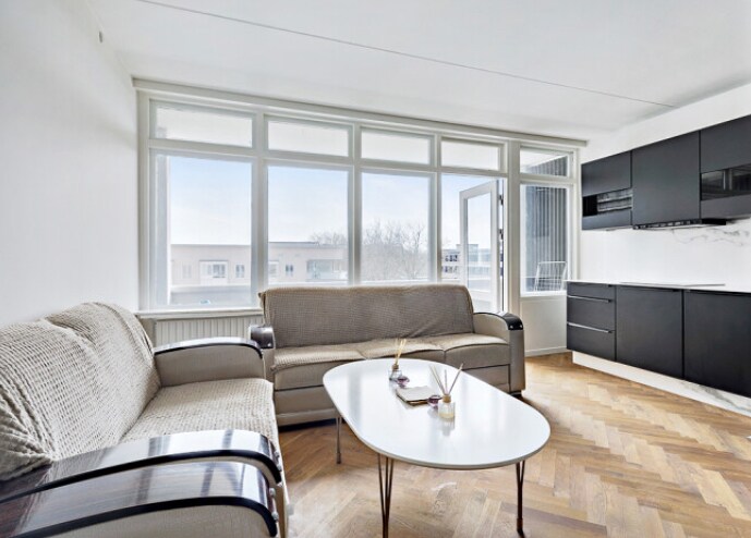 A bright living area features large windows that allow natural light to fill the space. Two comfortable sofas are positioned around a simple white coffee table. A modern kitchen is visible in the background, with dark cabinetry contrasted against light walls.