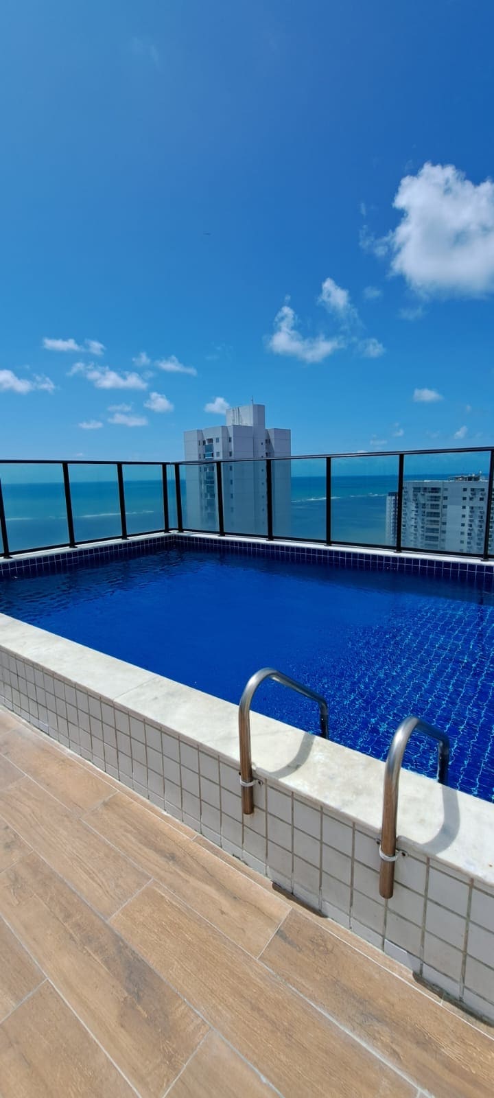 A rooftop pool is presented, featuring bright blue water and surrounded by glass railings. The expansive sky is visible overhead, with a few clouds drifting by. The city skyline and ocean can be seen in the background, providing a scenic and open atmosphere.