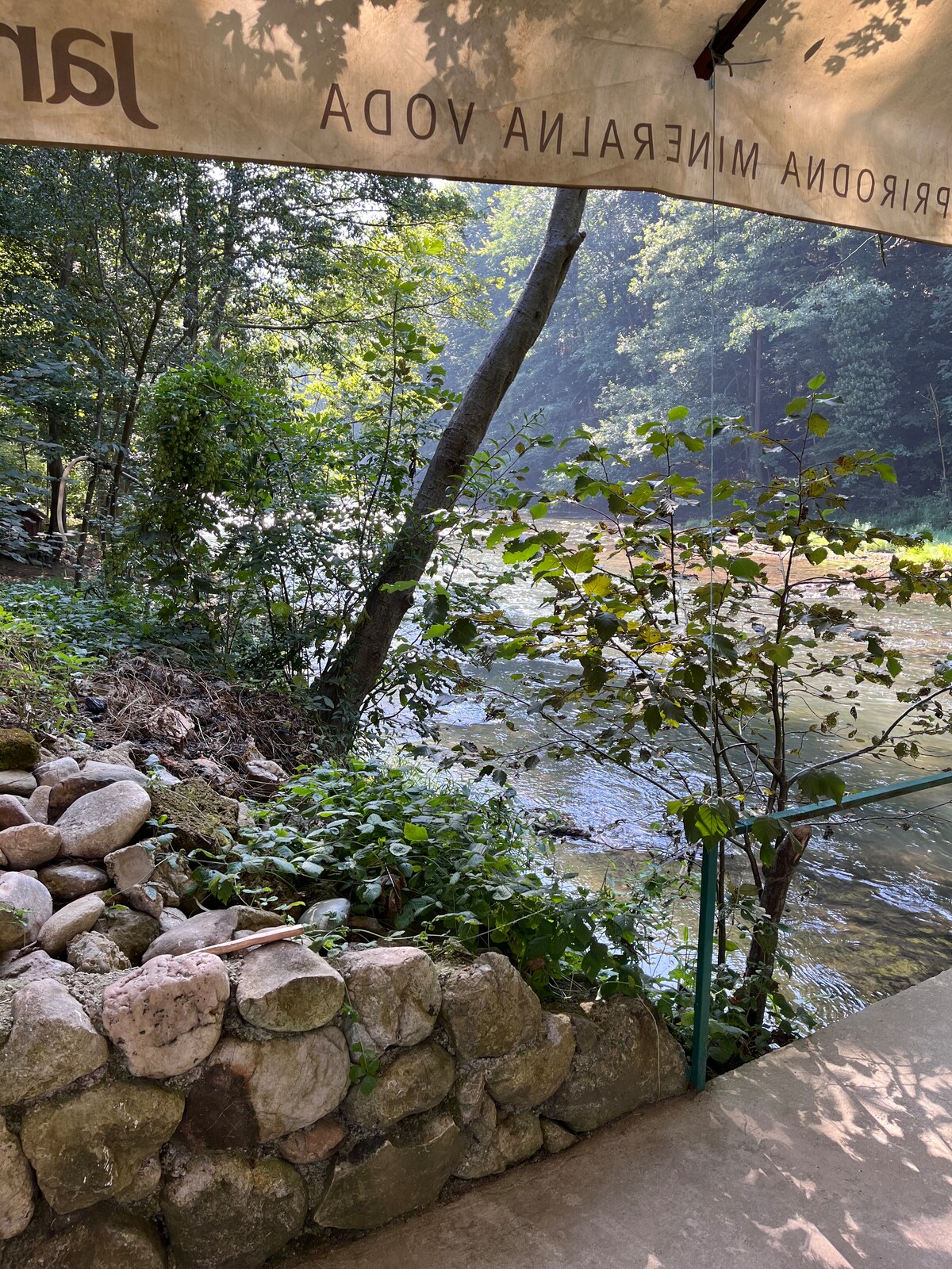 A peaceful view of a river flowing gently past lush greenery, with trees lining the bank. A stone wall and plants frame the scene, creating a serene outdoor setting, ideal for relaxation. Sunlight filters through the leaves, enhancing the tranquil atmosphere.