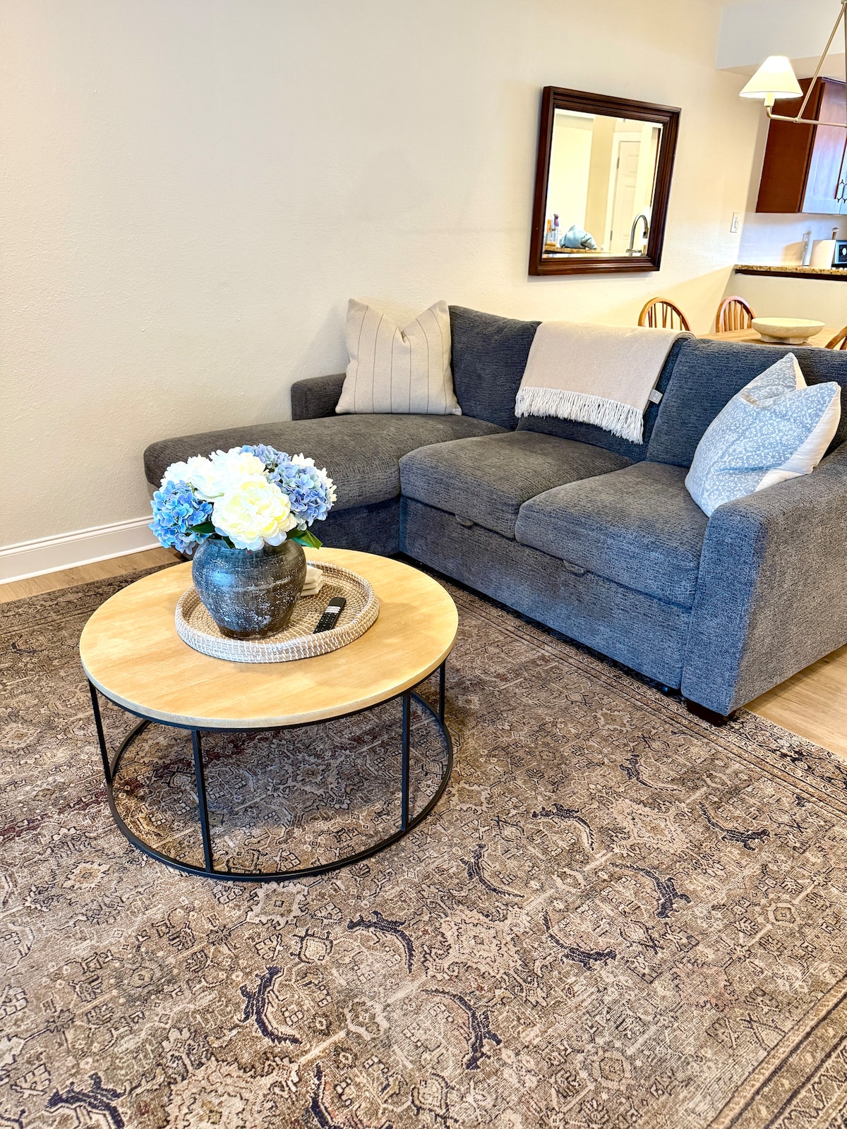 A cozy living area features a gray sectional couch adorned with decorative pillows. A round wood coffee table sits in front, holding a floral arrangement. Soft light filters in, highlighting the intricate patterns of the area rug beneath.