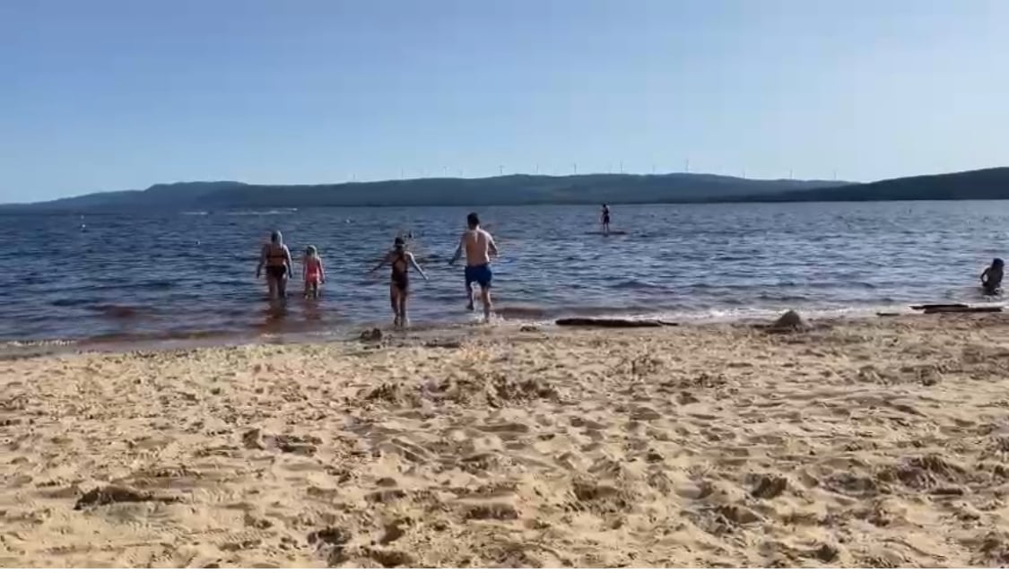 A sandy beach extends towards a calm lake, where several individuals are wading in the water. The backdrop features gentle hills and wind turbines in the distance, under a clear blue sky. Sunlight reflects off the water, creating a serene atmosphere.
