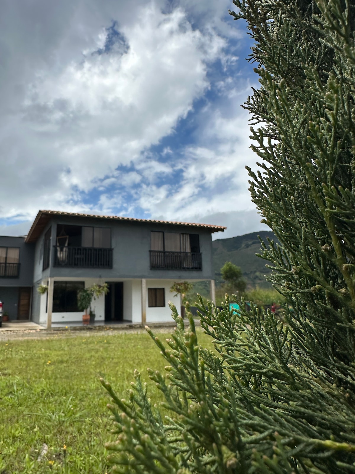 A two-story house is framed by greenery, showcasing a covered porch and balconies on the upper level. The cloudy sky adds texture above, while a grassy area leads to the entrance, creating a serene setting surrounded by nature.