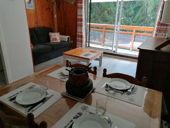 A dining table set with plates and glasses is positioned in the foreground, complemented by a black cookware pot. The living area features a dark sofa and a wooden coffee table. Large glass doors behind the seating area provide a view of lush greenery outside.
