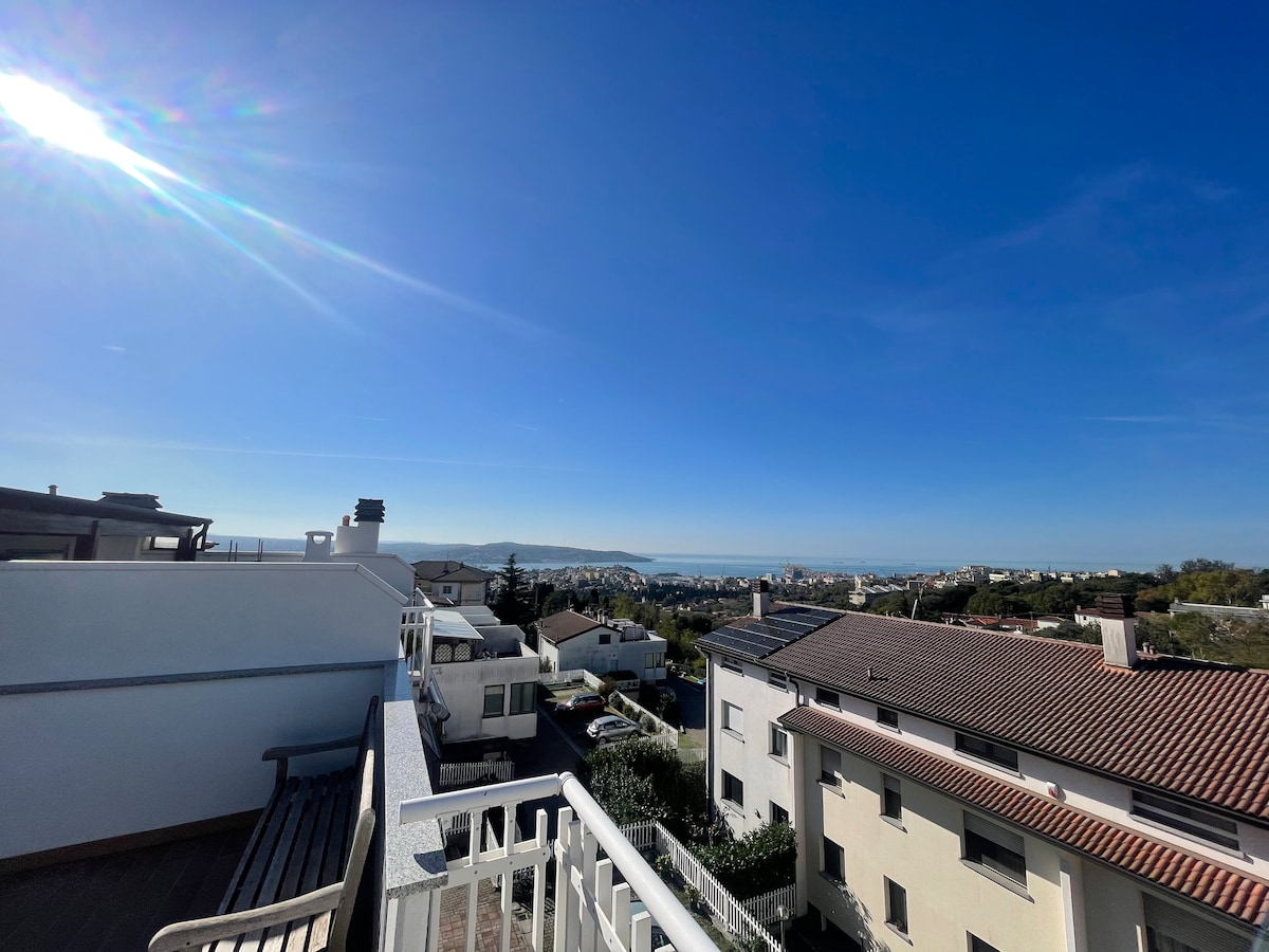 A panoramic view of the city and sea is presented from a terrace, with clear blue skies above. Nearby rooftops and greenery can be seen, creating a serene backdrop against the horizon.