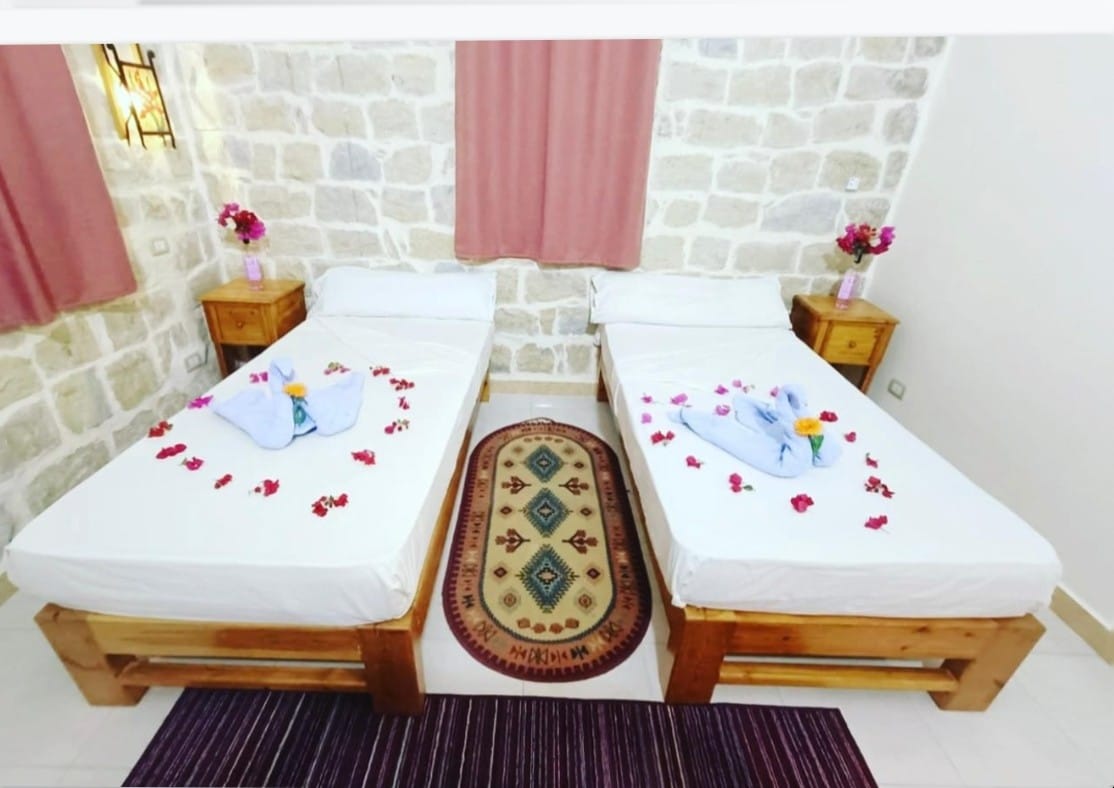 Two wooden beds are arranged in a cozy room, each adorned with floral arrangements and towels folded artistically. A colorful rug lies beneath, and a soft, neutral-toned wall made of stone adds texture to the space. Large windows are partially dressed with light fabric curtains.