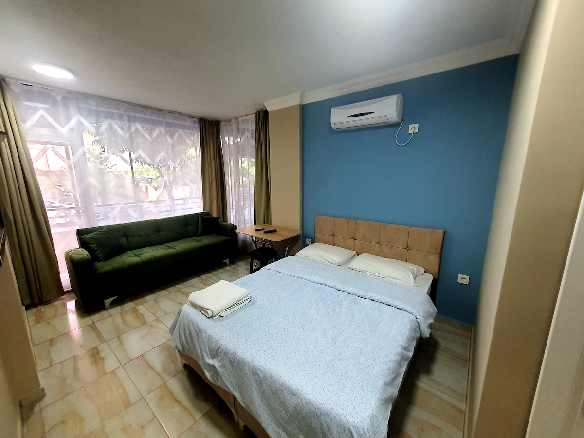 A cozy room features a spacious bed covered with a light blue blanket, positioned next to a comfortable sofa. A small table sits nearby, and large windows allow natural light to fill the space, framed by beige curtains.