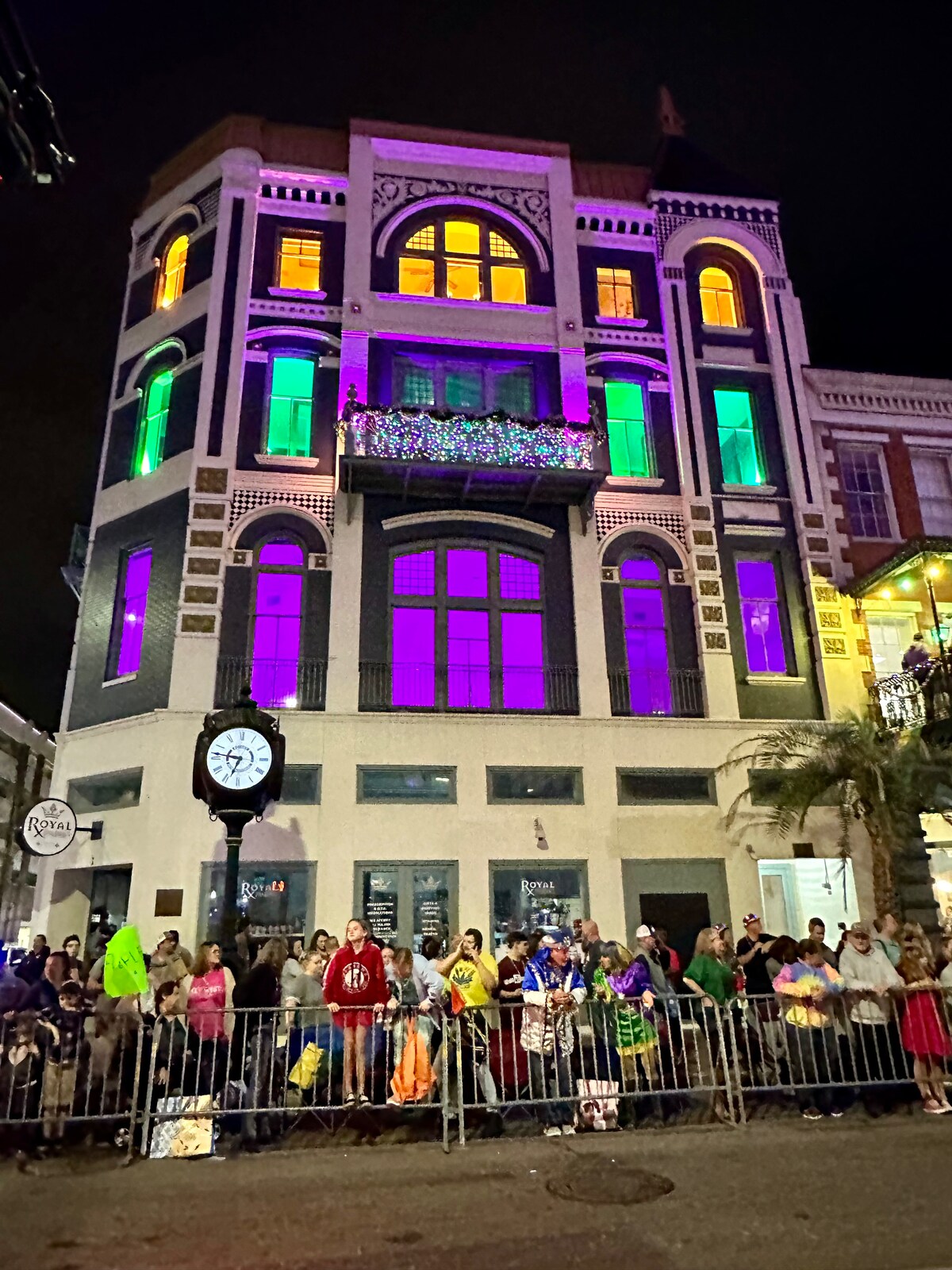 A vibrant building is showcased with purple, green, and yellow lights illuminating its architectural features. A large clock stands at the forefront, while a crowd gathers near a barrier at the base, adding to the lively atmosphere of the scene.