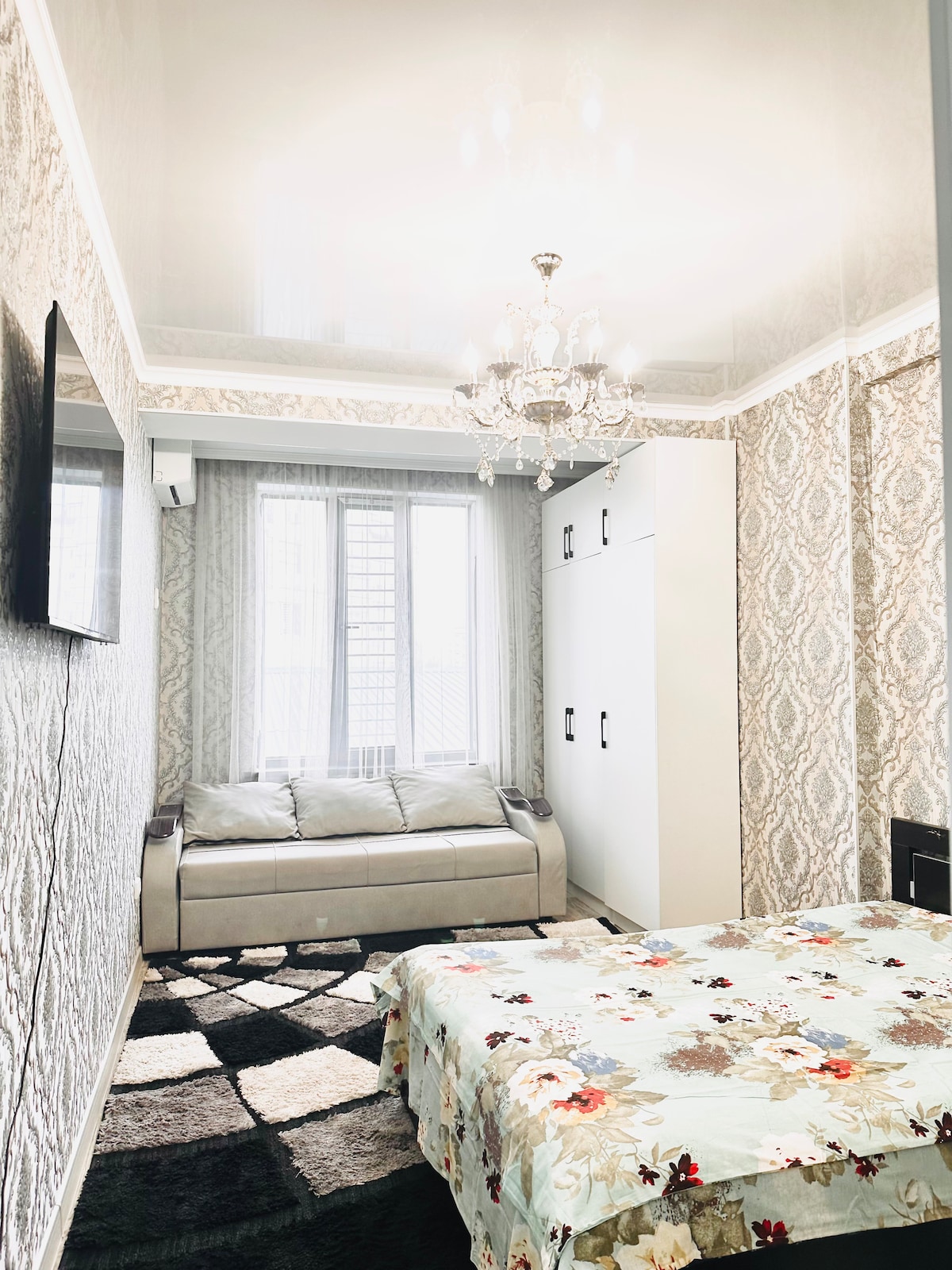A well-decorated room features a comfortable sleeping area with a floral-patterned blanket covering the spacious bed. A stylish chandelier hangs from the ceiling, and natural light filters through the large window. A sleek wardrobe and a cozy sofa are positioned against the patterned wallpaper.