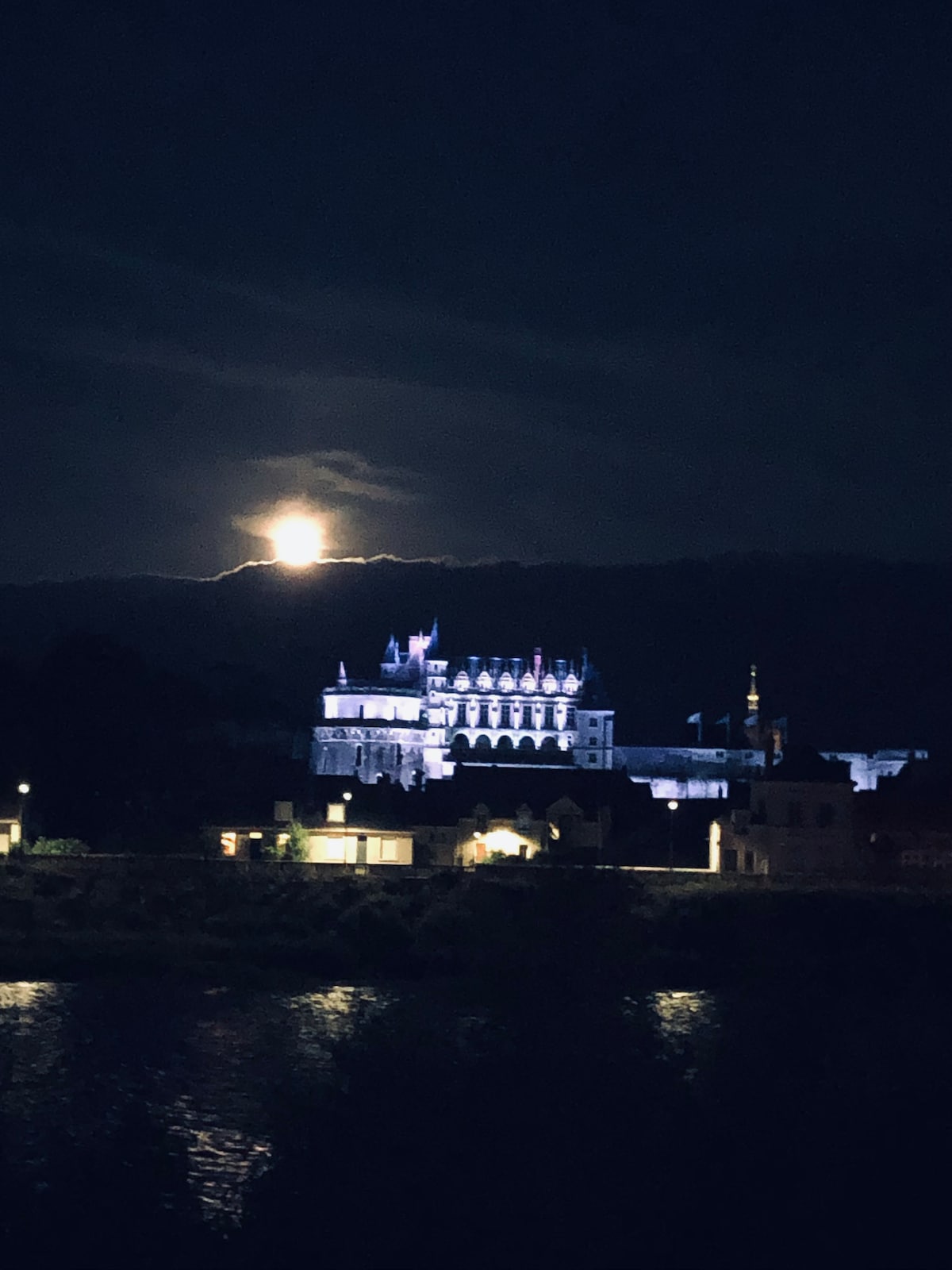 A moonlit view of a historical castle, with illuminated architectural features standing out against a dark sky. The still water in the foreground reflects the lights and silhouette of the castle, highlighting its grandeur in the night.