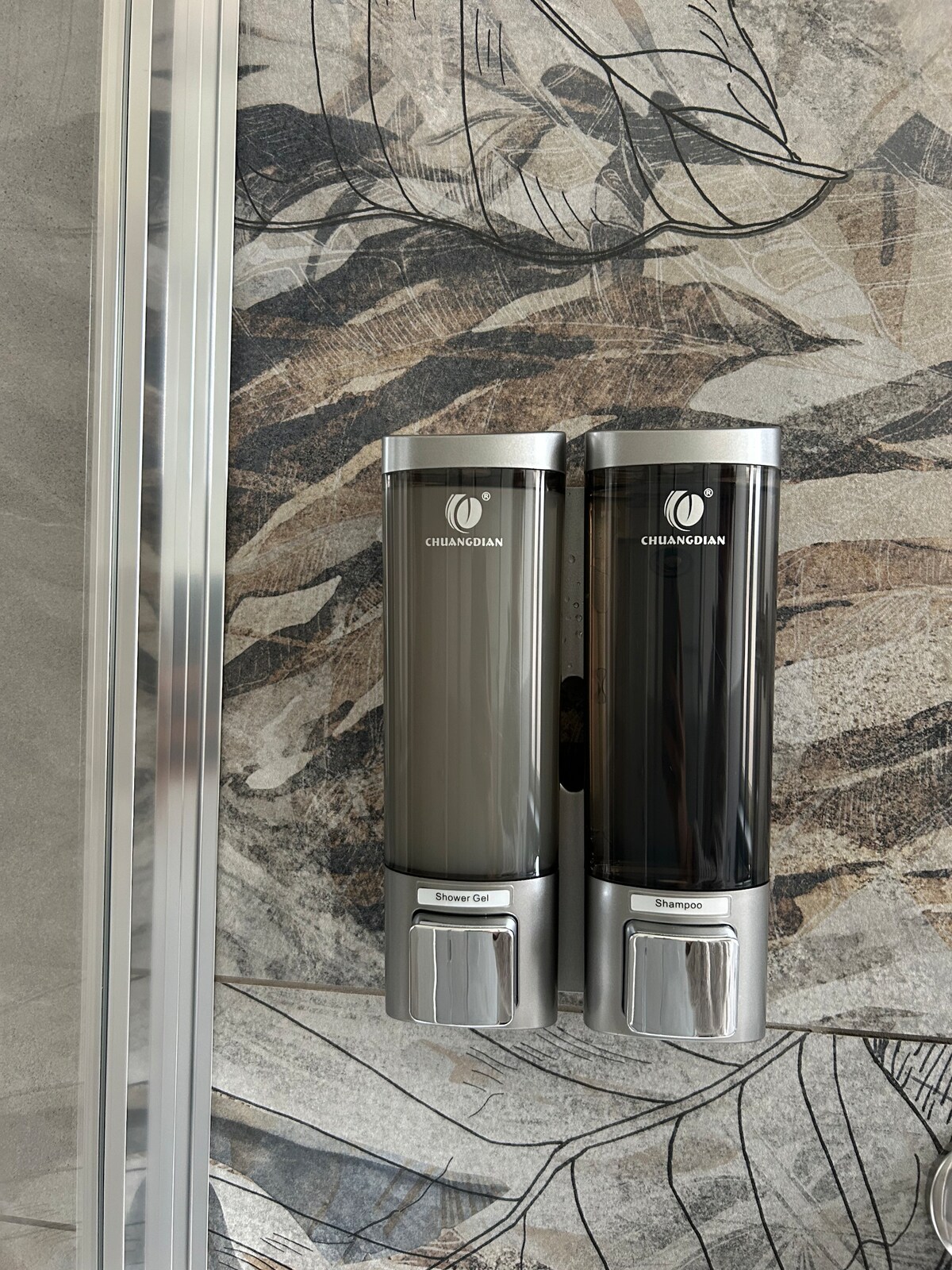 Two sleek dispensers are mounted on a textured wall with leaf motifs. One dispenser contains body wash, and the other holds shampoo, both labeled for easy identification.