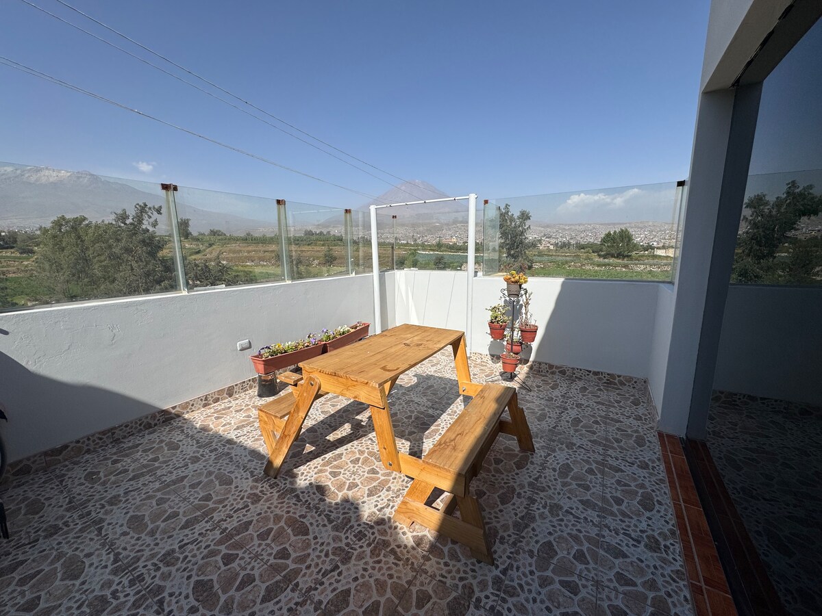 An outdoor terrace features a wooden table and bench set, surrounded by potted plants. The area offers expansive views of the landscape and distant volcanoes, with sunlight illuminating the stone floor. Glass panels provide safety while maintaining an open feel.