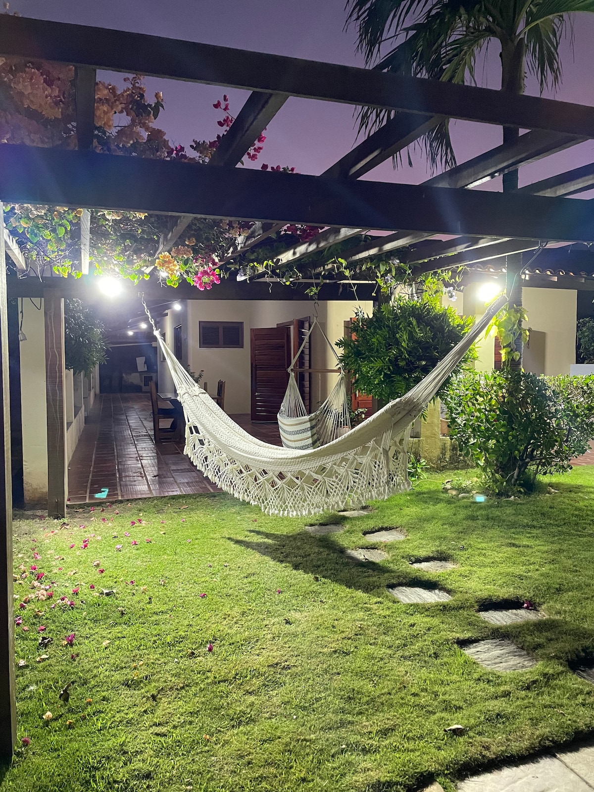 A serene outdoor area is presented, featuring a hanging hammock beneath a wooden pergola. Soft lighting enhances the garden's greenery, while scattered flowers grace the grassy lawn. Pathway stones lead through the space, creating an inviting atmosphere for relaxation.