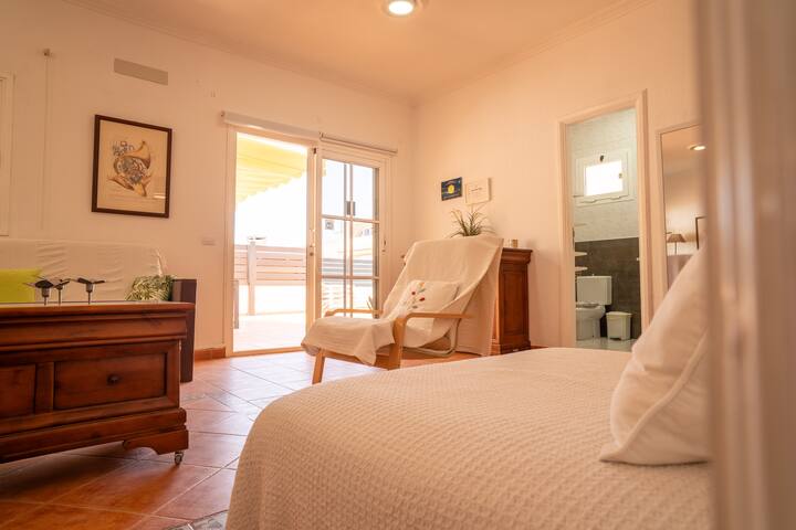 Estancia luminosa con cama, butaca y salida a la terraza, junto al baño: espacio versátil y tranquilo.