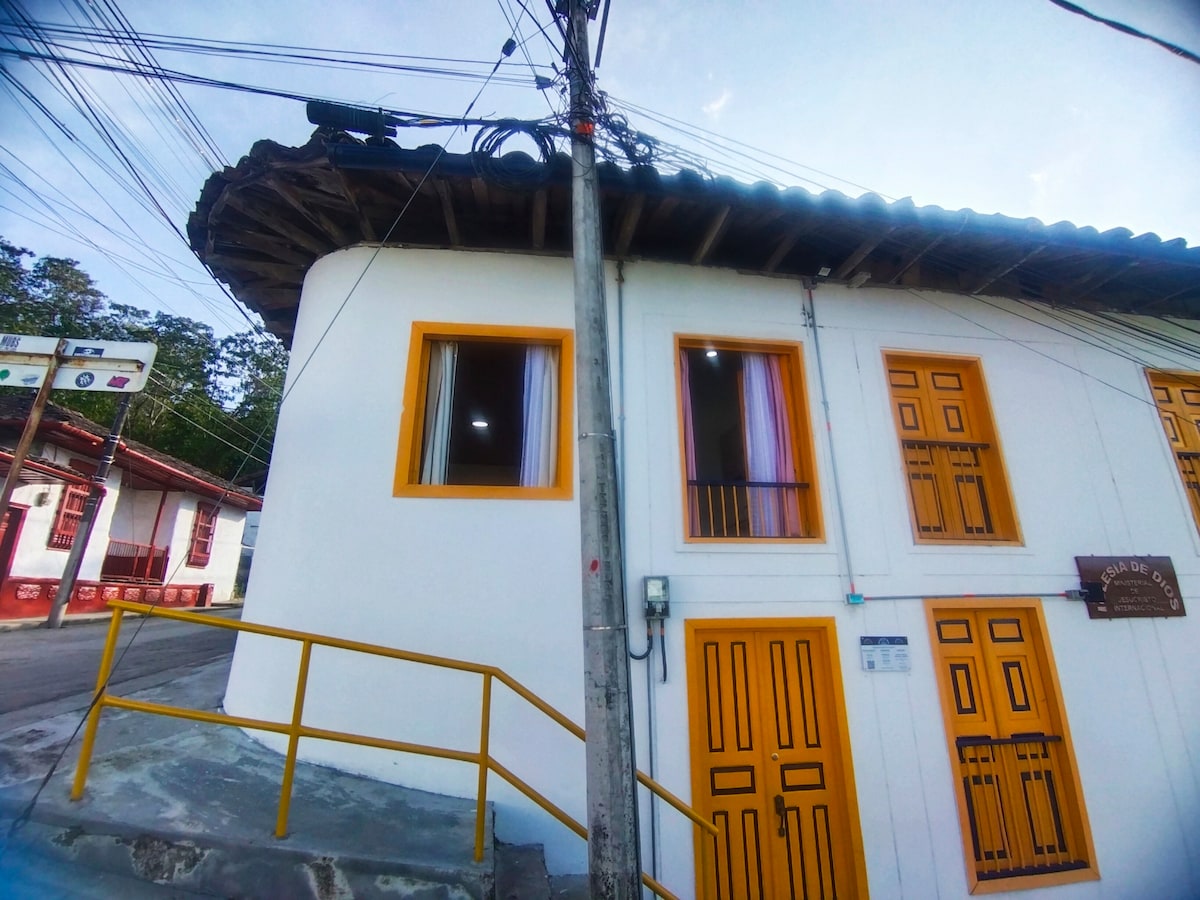 The exterior of a colorful building features white walls and vibrant yellow doors, showcasing large windows with light curtains. A gently sloped roof adds to its charm, while utility poles and power lines are visible in the background.