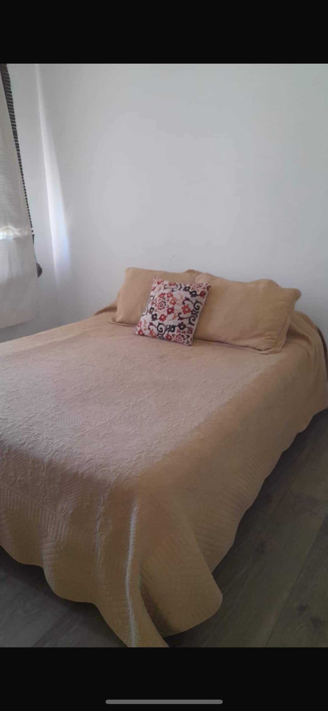A spacious bed is dressed in a light-colored coverlet, complemented by decorative pillows. Natural light filters through a nearby window, which is dressed with sheer curtains, creating a calm sleeping area.