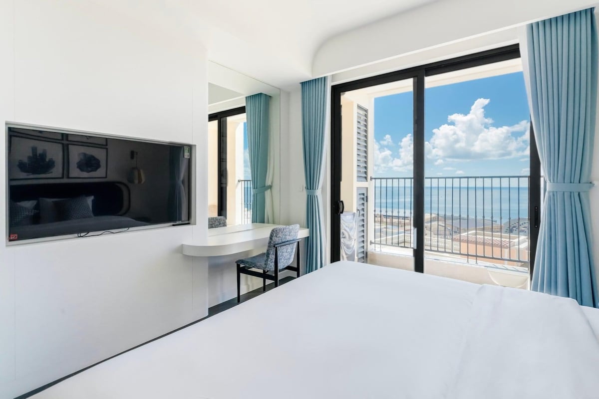 A bright bedroom is featured with a spacious bed and large windows, offering a view of the sea and sky. A flat-screen television is mounted on the wall, and a desk with a chair is positioned beside it, enhancing the functional layout.