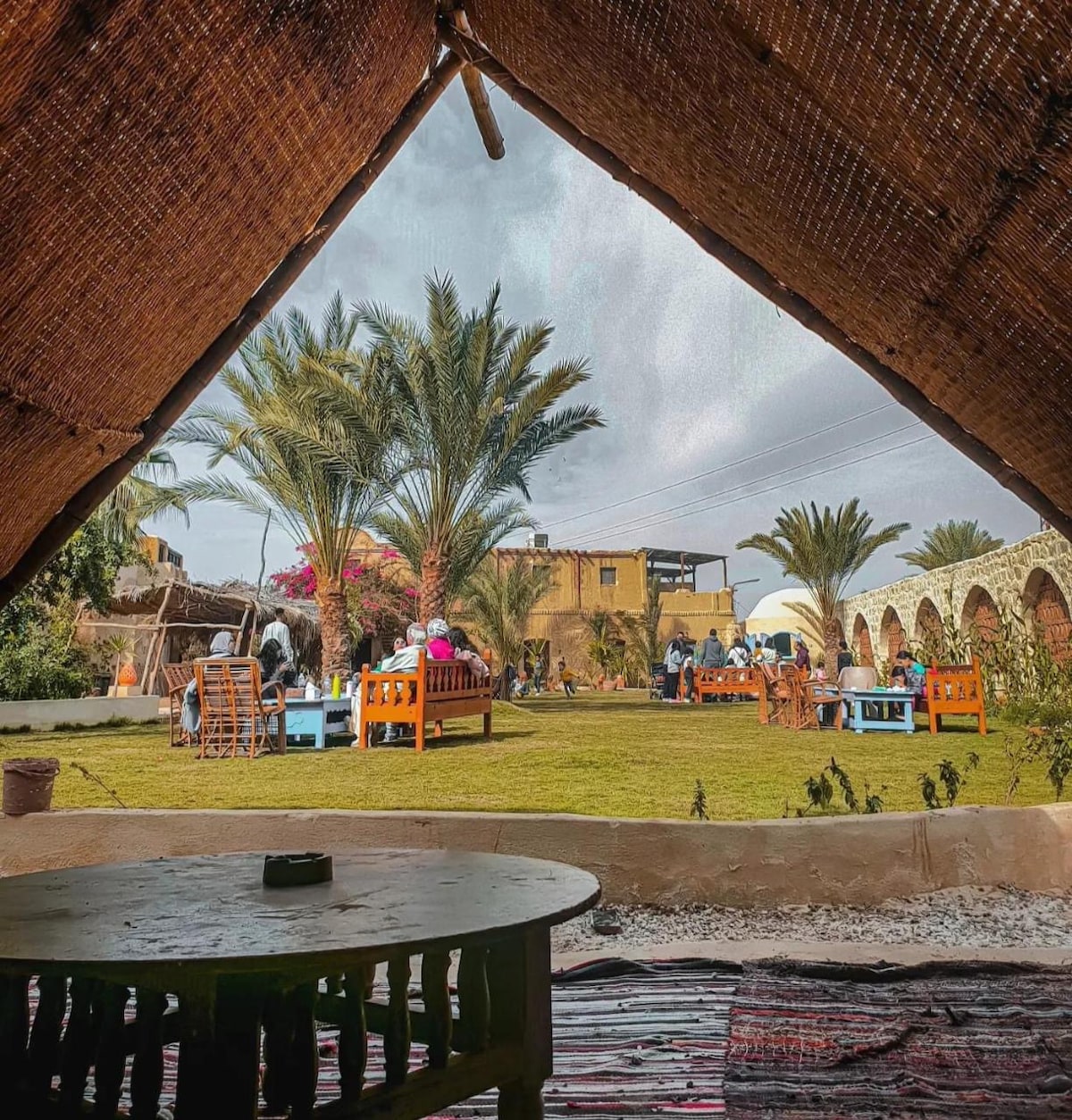 An outdoor area is framed by a woven palm roof, showcasing a well-kept lawn dotted with wooden furniture. Palm trees provide shade, and colorful flowers add visual interest. Guests can be seen enjoying the relaxed atmosphere within this inviting space.