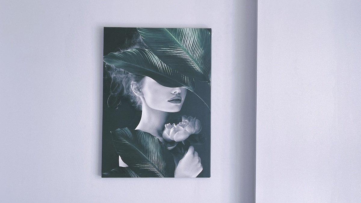 A wall-mounted artwork features a blend of large green leaves and a white rose. The composition includes a central area that appears intentionally obscured, emphasizing the surrounding botanical elements against a soft, light-colored wall.