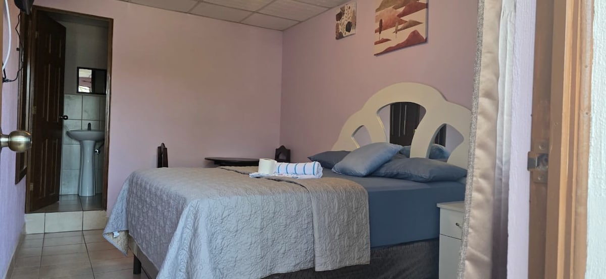 A calming sleeping area is presented with a spacious bed adorned with blue pillows and a neatly folded towel. Soft purple walls create a serene atmosphere. An ensuite bathroom is visible through an open door, featuring white fixtures and simple decor.